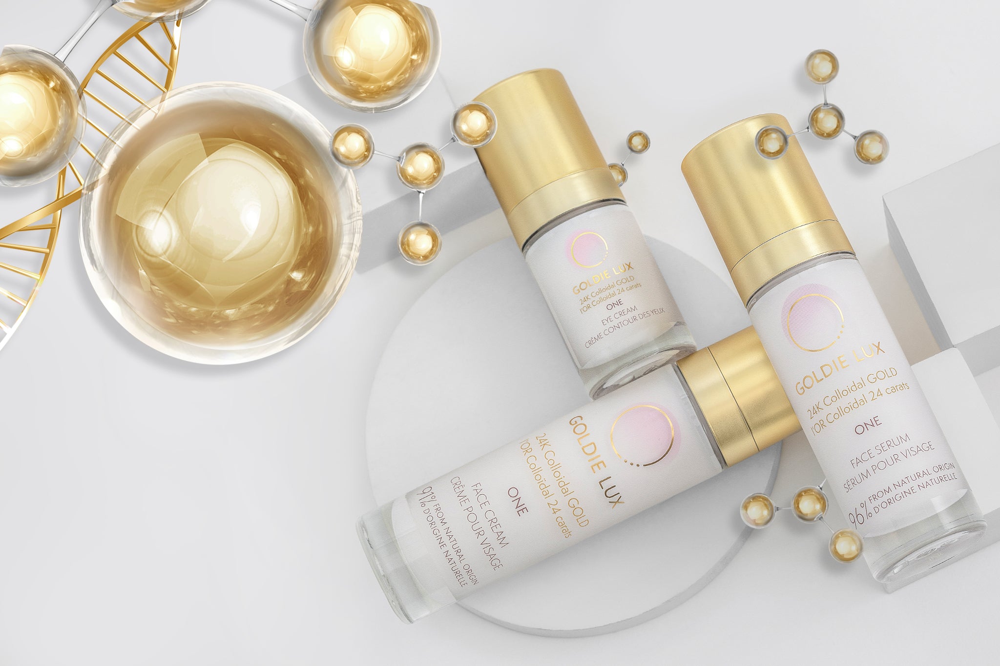 Goldie Lux | 24 Karat Colloidal Gold One Collection featuring face serum, eye cream, and face cream glass bottles with gold caps, surrounded by gold molecular structures.