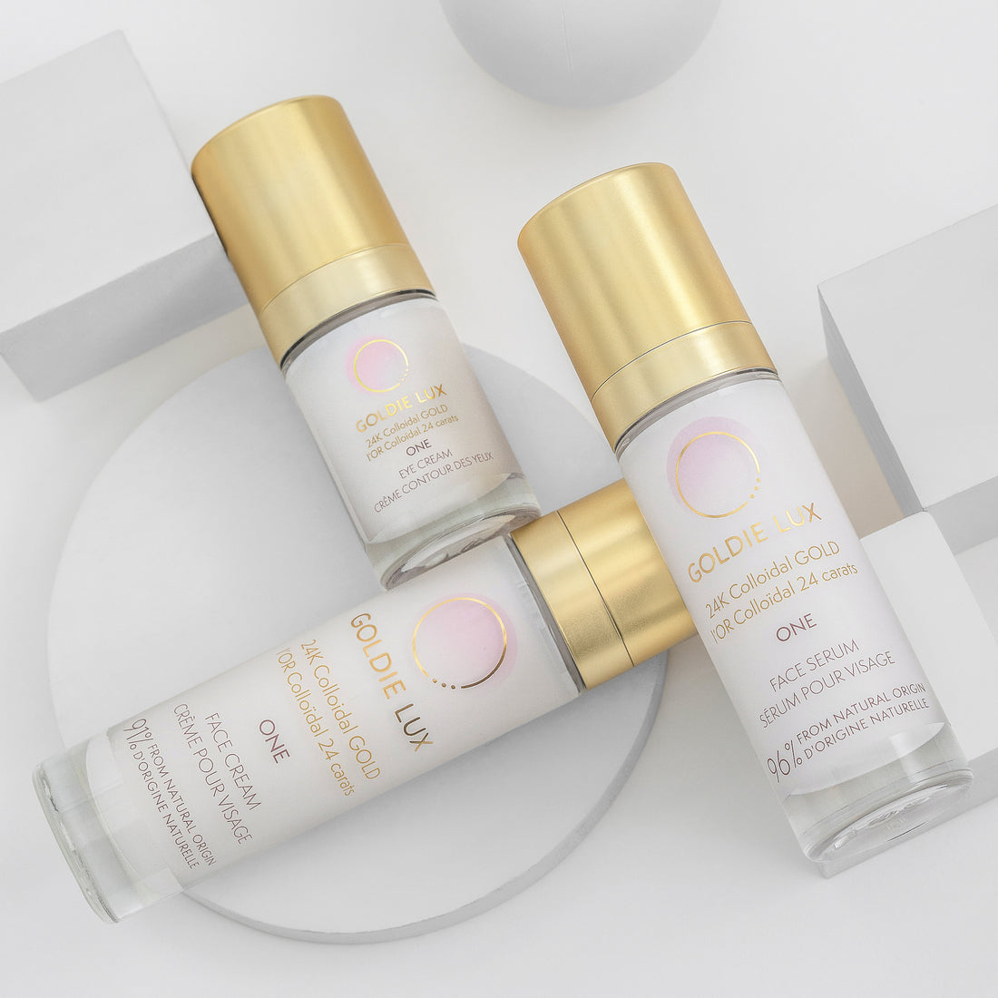 Goldie Lux | 24 Karat Colloidal Gold One Collection featuring face serum, eye cream, and face cream glass bottles with gold caps on white background.